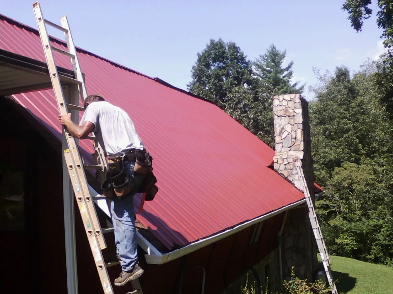 Roofer on a ladder beside a red metal roof and stone chimney — Soffit & Fascia Repair by Roofing Brothers in Riceboro